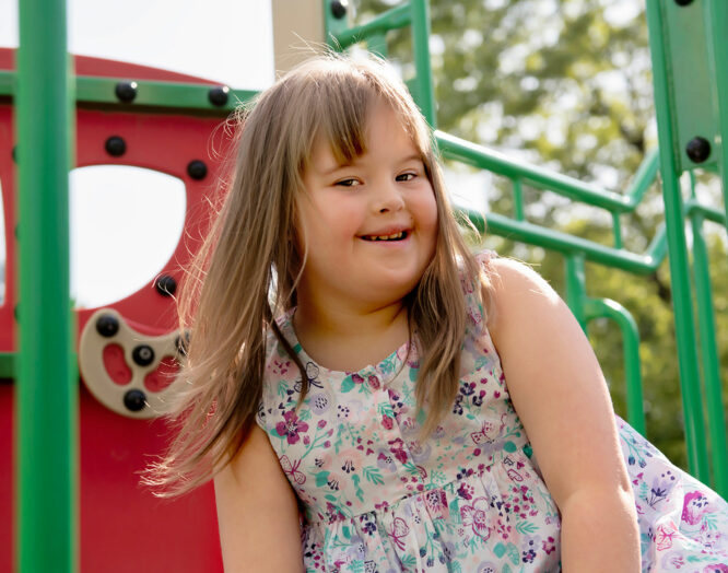 Ein Mädchen mit Trisomie 21 steht lächelnd vor einem Spielplatz.