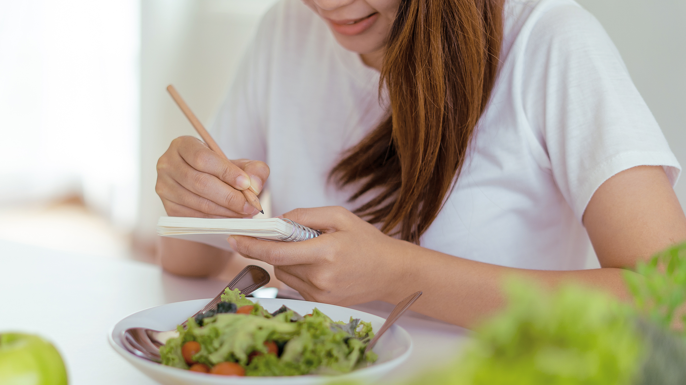 Eine Frau sitzt vor einem Teller Salat und macht Notizen in ein kleines Notizheft